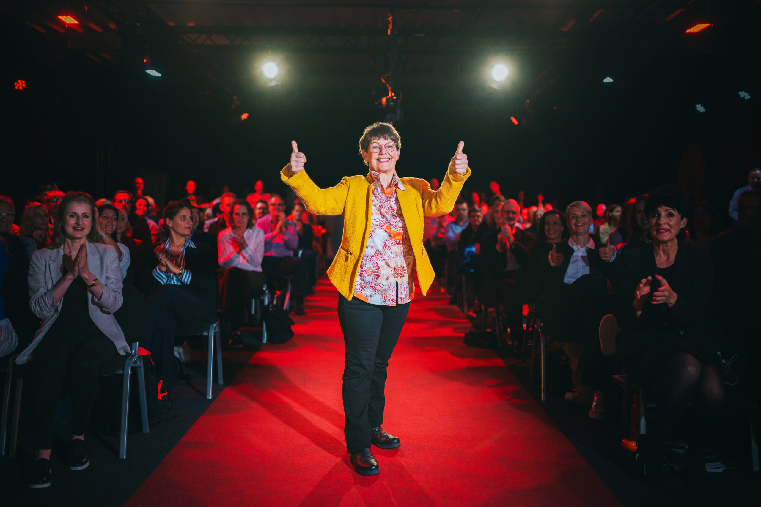 Stella Reichmann steht freudestrahlend im Mittelgang eines mit Zuschauern besetzten Saales. Sie bekommt tosenden Applaus und hält beide Daumen zum Zeichen ihrer Freude in die Luft. Sie trägt eine schwarze Hose, gelben Blazer und eine orange gemusterte Bluse.