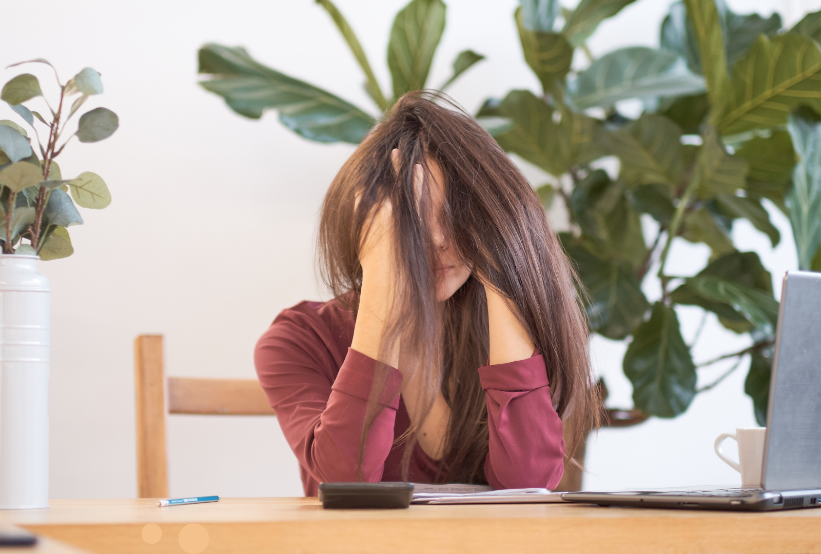 Eine Frau sitzt erschöpft an einem Schreibtisch, den Kopf in die Hände gestützt. Ihr Gesicht ist teilweise von ihren langen Haaren verdeckt. Auf dem Tisch liegen Unterlagen, ein Handy und ein geöffneter Laptop. Im Hintergrund stehen große grüne Zimmerpflanzen. Die Szene vermittelt Frust, Überforderung oder Erschöpfung im Arbeitskontext.