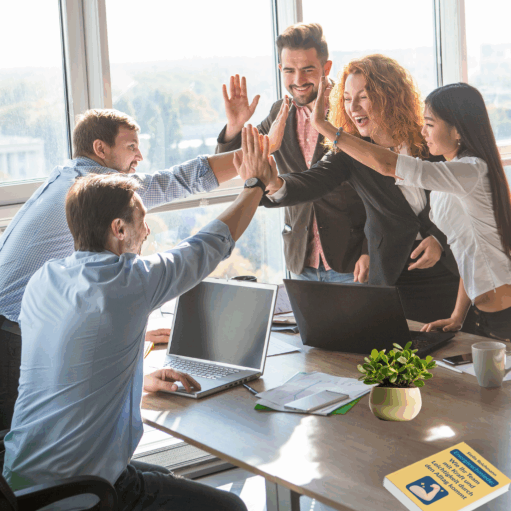 Fünf beruflich gekleidete Menschen stehen lachend um einen Tisch in einem modernen Büro mit großen Fenstern. Sie geben sich alle gleichzeitig ein High-Five in der Mitte – ein Ausdruck von Teamgeist und gemeinsamer Freude. Auf dem Tisch liegen Laptops, Notizblöcke, eine Tasse, eine grüne Zimmerpflanze und ein gelbes Buch mit dem Titel: "Warum Teams mit Freude leichter führen, denken und arbeiten lernen" von Stella Reichmann. Im Hintergrund sieht man eine helle, sonnige Landschaft durch die Fenster. Die Stimmung ist dynamisch, motivierend und positiv.